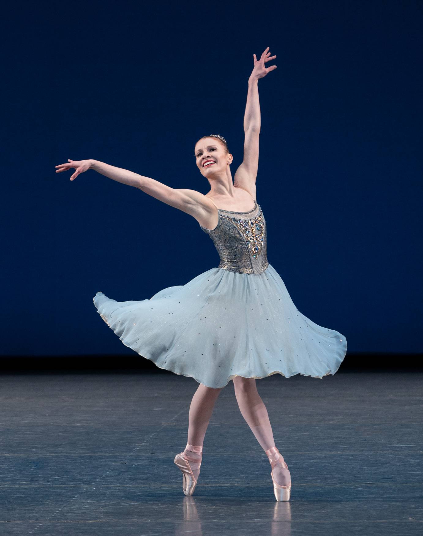 A woman poses en pointe with her legs crossed, her arms amplified